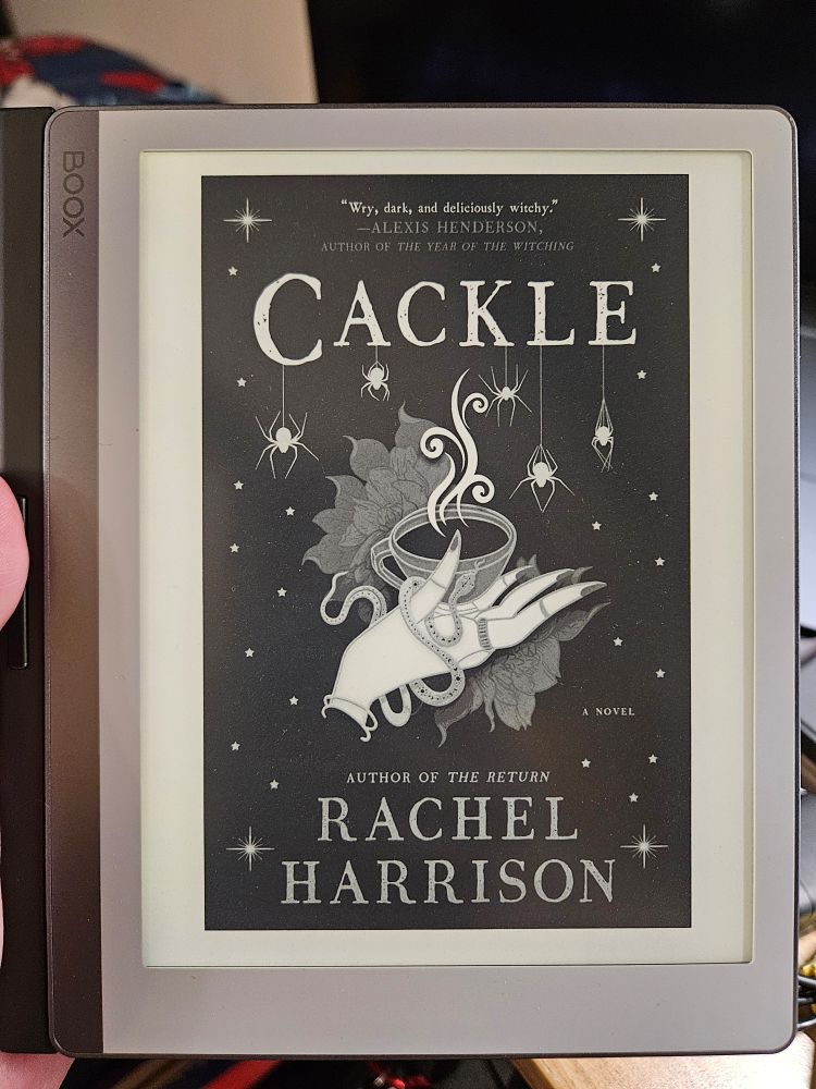 Black and white cover of Cackle by Rachel Harrison. A singular hand with manicured nails is holding a teacup with steam rising from the top. A snake is wrapped around the hand. White spiders are hanging from a black spotted background.