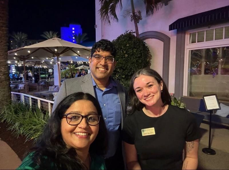 Three Flaum faculty and or alumni posing for a group selfie, two women one man, at night on the back patio of a restaurant. 