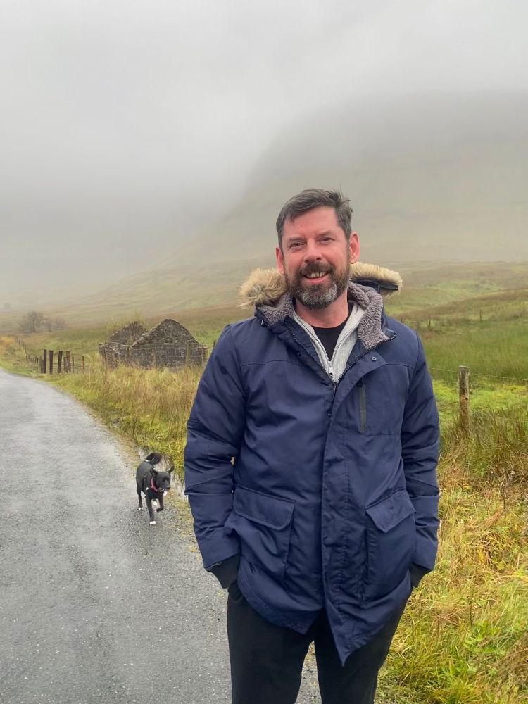 A picture of me in a heavy blue jacket standing on a rural road known as the Gleniff Horseshoe in County Sligo.
Behind my right shoulder is a small ruined homestead. Marley, my black dog is trotting down the road behind me.
