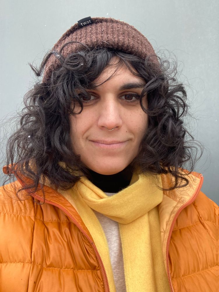 Esme is smiling like 🙂 , she’s wearing an orange coat with a yellow scarf and a brown beanie that says “twat”