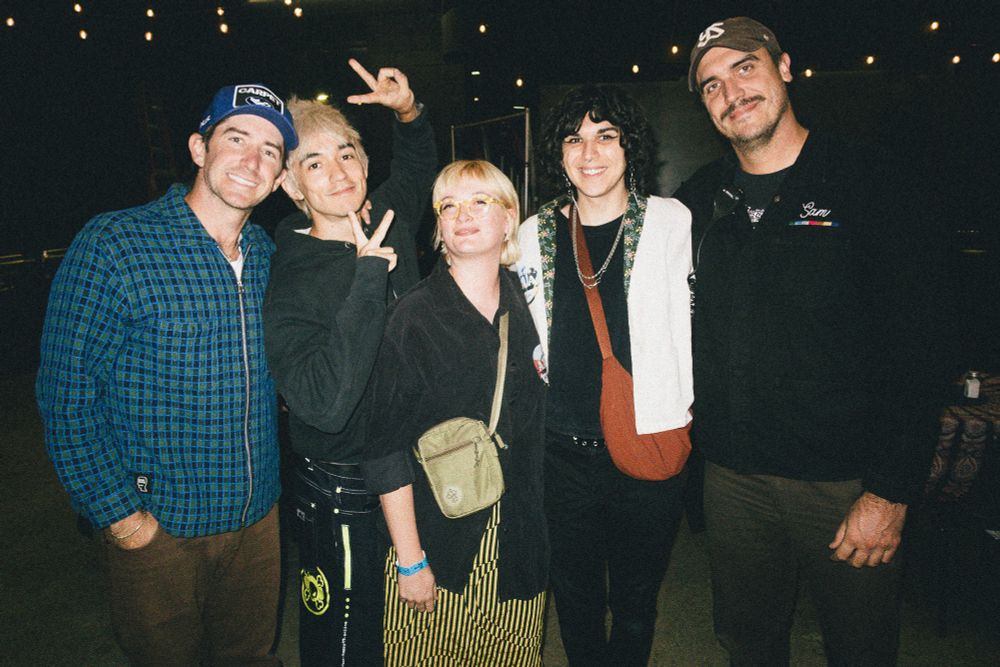 a photo of five people, from left to right: Pat and Daniel from turnstile, Steph, Esme, and Sam. They’re smiling and huddled in, Daniel is posing with his fingers like some cool anime character. Photo taken by legend Atiba 