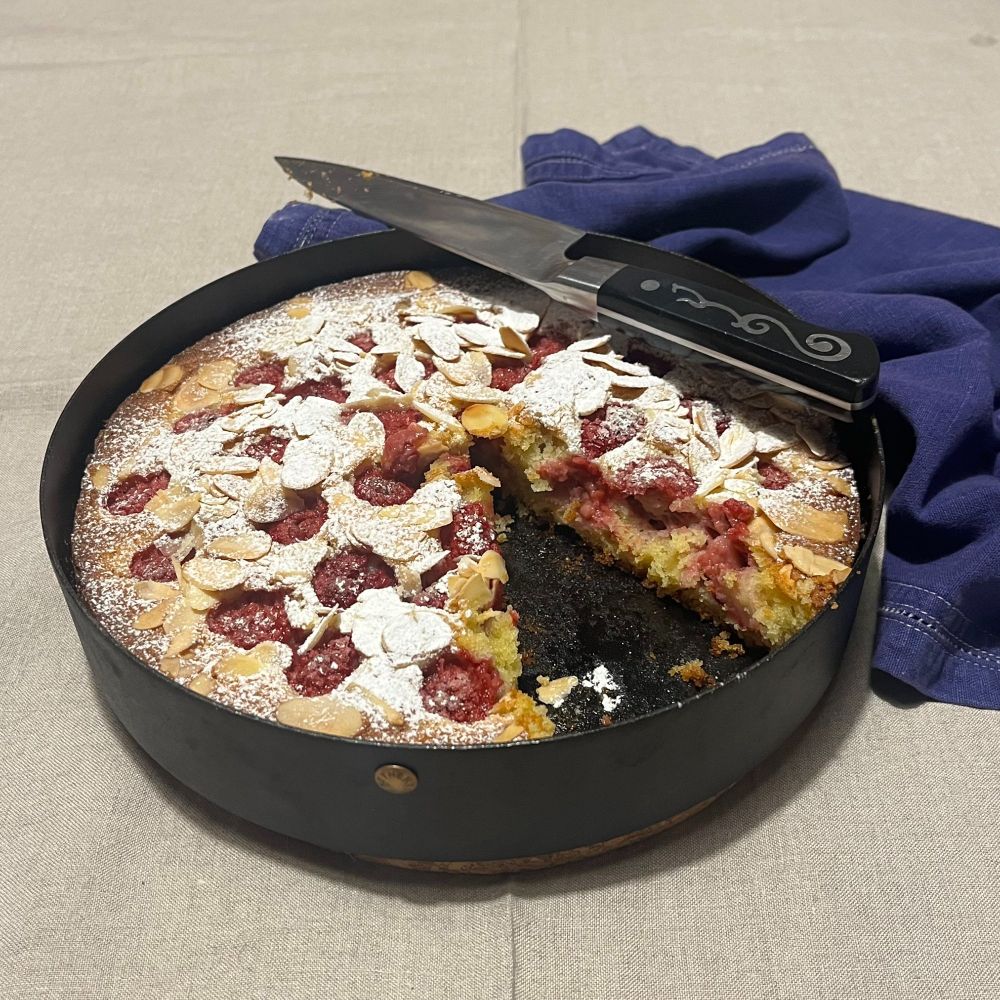 A raspberry frangipane tart, with one slice removed, in a Netherton Foundry cake tin, with an IOShen knife and a dark blue rumpled napkin