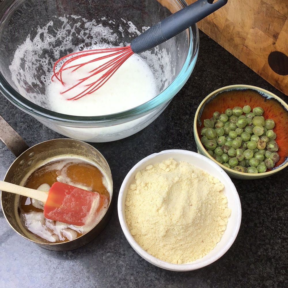 A mise en place scene of whisked egg whites i a glass bowl, a bowl full of ground almonds, flour and icing sugar, a Sytch farm bowl of gooseberries and a Netherton Foundry copper pan containing melted butter