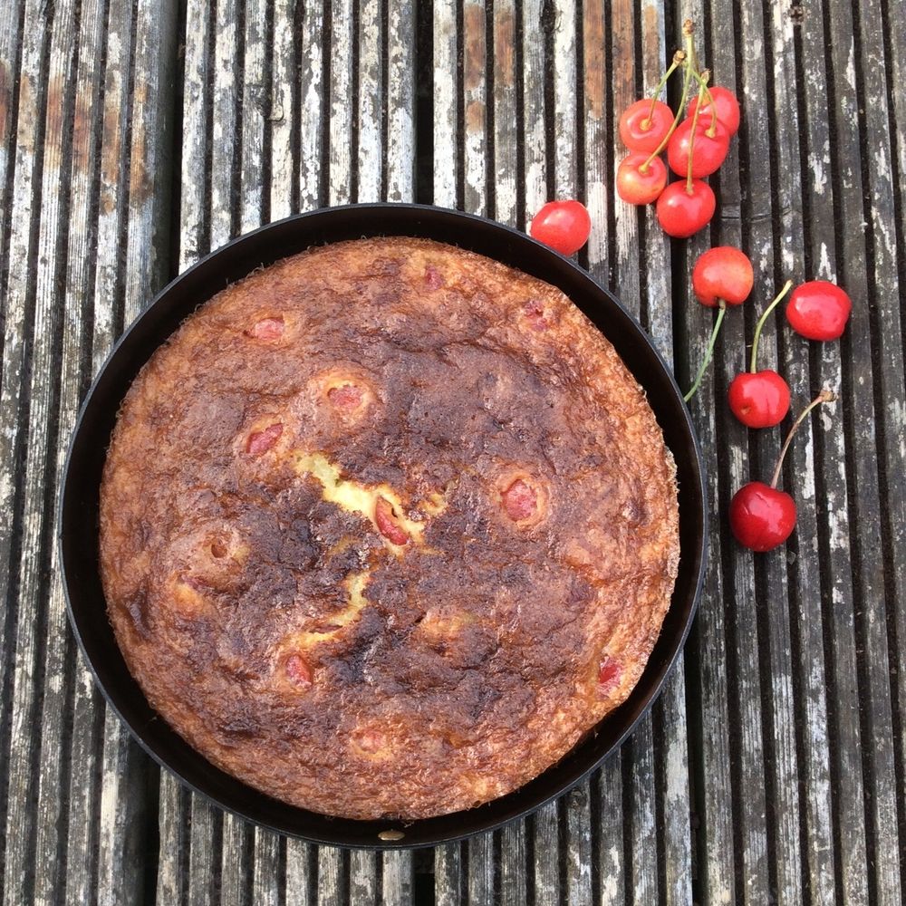 A baked cherry and almond batter pudding in a Netherton Foundry cake tin on a wooden surface with a scattering of fresh cherries alongside