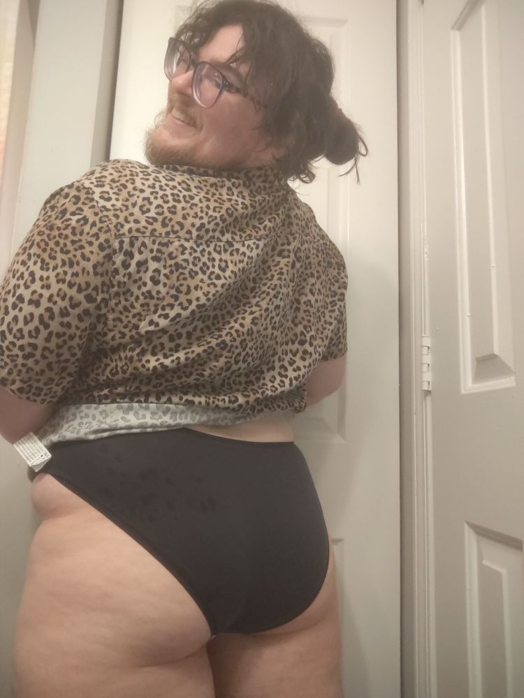 TJ smiling over his shoulder. His back is to the camera, showing his thick thighs and butt. He's wearing black bikini briefs and a cheetah print shirt