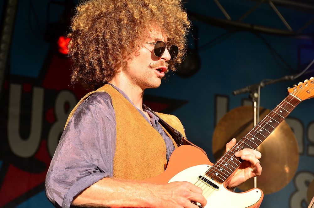 Ein Gitarrist mit blonden Locken einer braunen Weste einem lila Hemd und einer Sonnenbrille und da hinter unser Banner