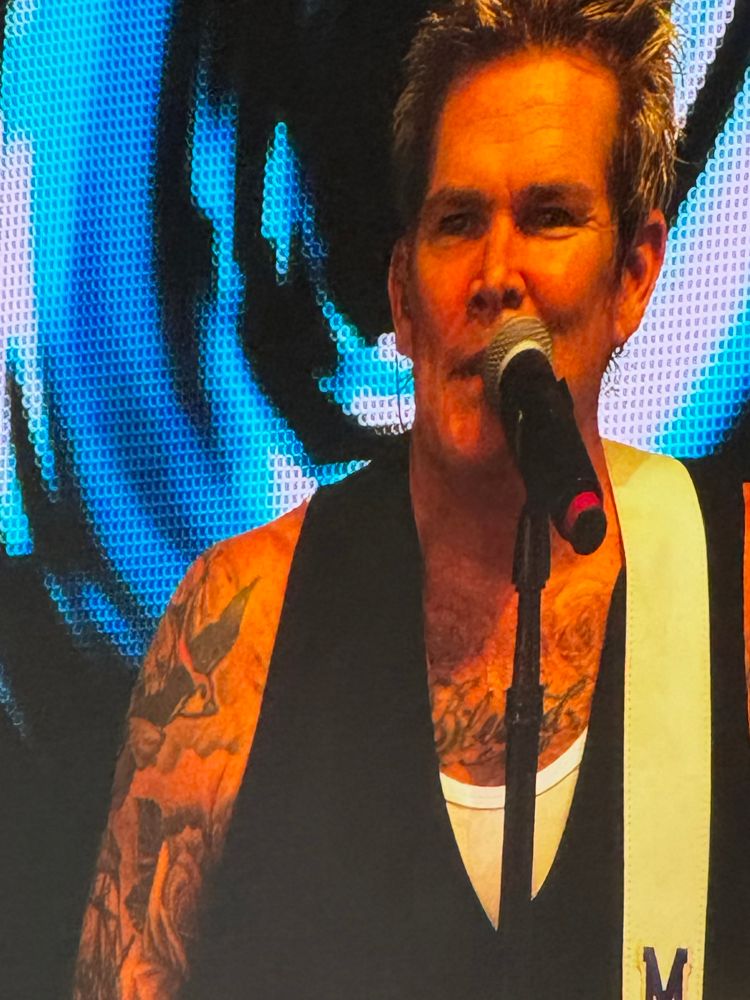 Photo of an aging rockstar on a Jumbotron screen. Spiky, bleached blonde hair. High forehead. Impressive cheekbones. He’s wearing a black waistcoat over a white singlet. Exposed arms and chest are covered in tattoos. White guitar strap on his left shoulder.