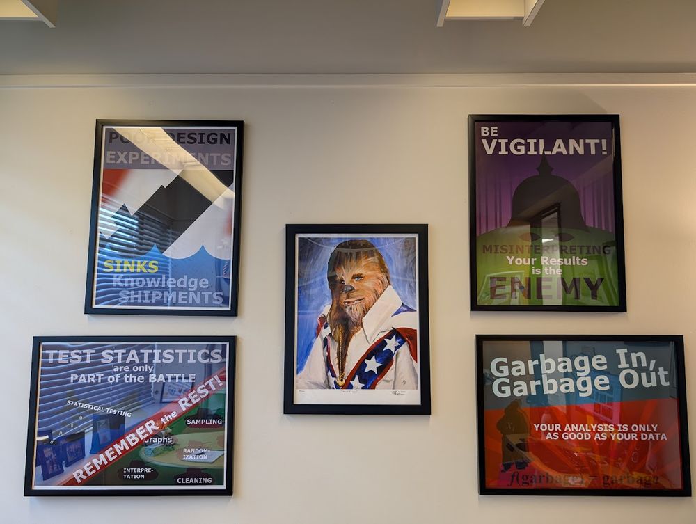 Photo of an office wall with four World War II style propaganda posters: 1) Poor design of experiments sinks knowledge shipments; 2) Test statistics are part of the battle. Remember the rest! statistical testing, graphs, sampling, randomization, interpretation, cleaning; 3) Be vigilant! Misinterpreting your results is the enemy; 4) Garbage in, garbage out: Your analysis is only as good as your data. In the center is an art print of Chewbacca dressed as Evel Knievel