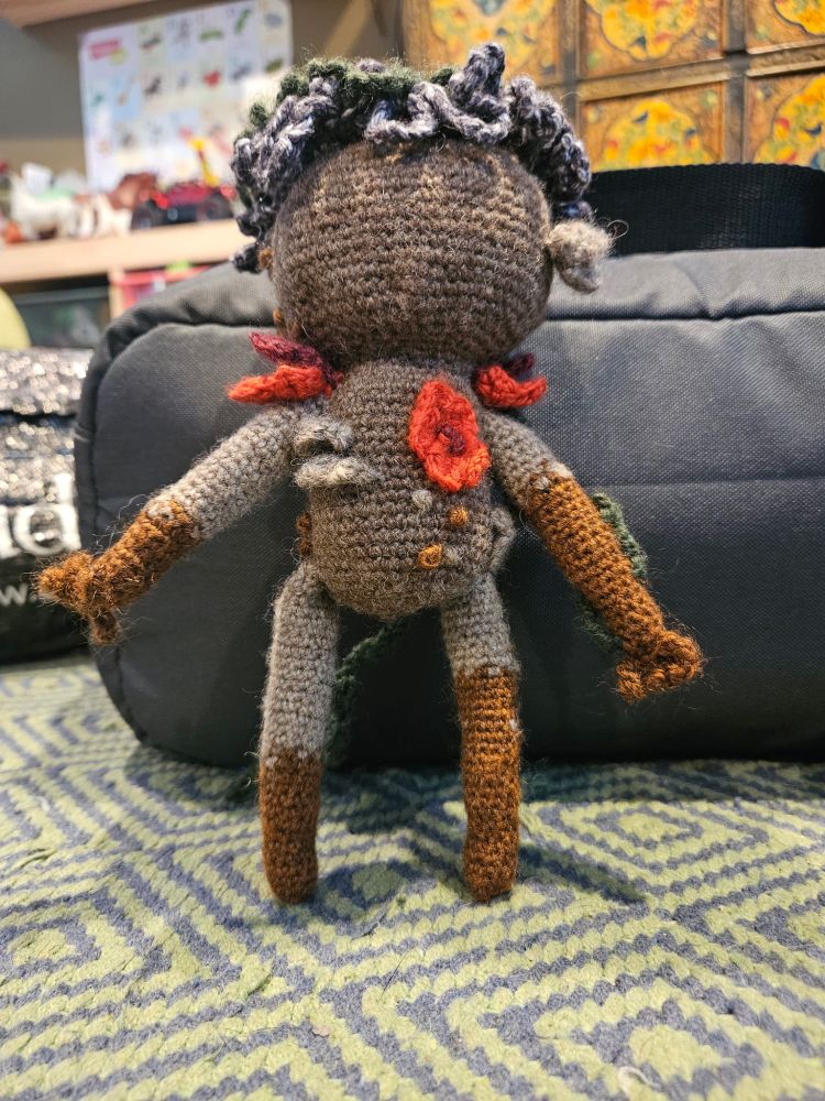 Picture of a crochet Adne Sadeh vegetable man standing up, with it's back toward the camera, showing off flowers and (I think) mushrooms