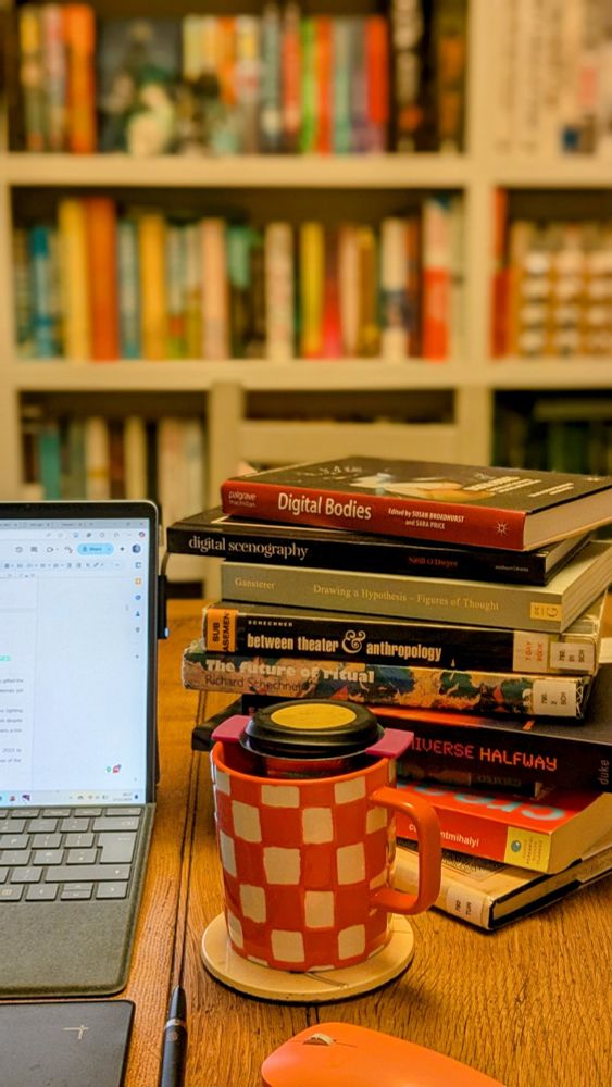 The focus of the photo is a large orange + white checked mug of tea, sat on a wooden table top.
Around it on the table are a pink computer mouse, the visible edge of a laptop and graphics tablet and a stack of books on digital theatre, ritual, creativity and entanglement.
In the background is an out of focus colourful bookshelf.