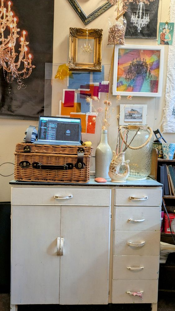 Photo shows a vintage white cupboard with pale blue top in front of a wall covered in colourful pictures, prints and photos of chandeliers. It is a cluttered, busy space.
On the cupboard sits a picnic hamper with a laptop on top.