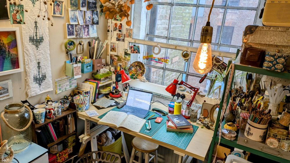 Photo of a very pretty, cosy and cluttered desk in an art studio. The wall to the left is covered in pictures and a set of shelves to the right is packed with art supplies and pots of pencils and paintbrushes.
On the desk is a laptop and open notebooks.