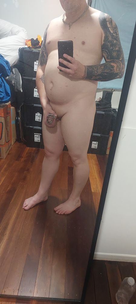Man with tattoos in chastity, holding his Master's cock. 