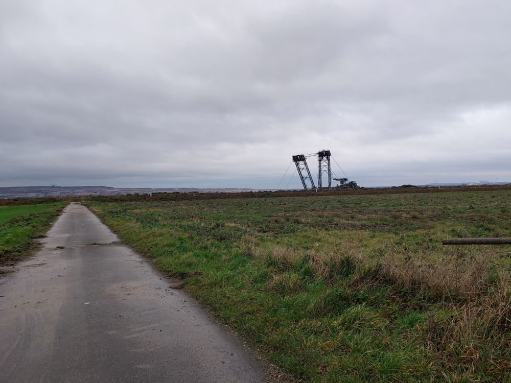 Grün/braunes Feld vor dem Wall dahinter steht das Monster, der kohlebagger. Geteerter Feldweg der zum Tagebauvorfeld führt