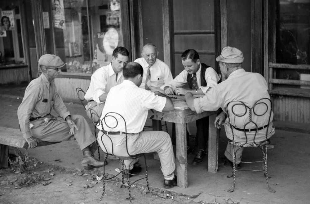 Drugstore dominoes: Four men mind their dominoes at a makeshift table outside a drugstore in Mississippi Delta, Mississippi, in October 1939. Two observers, one on a bench at the left and the other with a tie, wire-rim glasses and a pen in his shirt pocket, observe the play closely. The players’ metal chairs appear to be borrowed from a soda fountain in the drugstore. Marion Post Wolcott made the photograph.