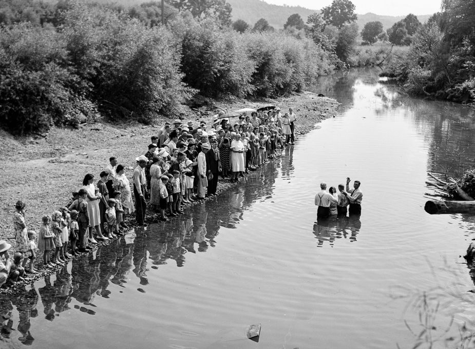 Praise the Lord: Congregants of the Primitive Baptist Church line the left bank of a creek as the pastor and another church official prepare to conduct a baptismal ceremony in the creek. They stand in knee-deep water; the pastor has his right arm raised as he prays over the couple to be baptized by submersion. Marion Post Wolcott photographed the scene in August 1940 near Morehead, Kentucky.