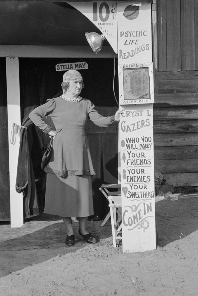 At the “Temple of Knowledge” booth at the Louisiana State Fair in November 1938, Russell Lee immortalized fortune teller Stella May, identified by the small sign over her head. She wears a gypsy head scarf and holds a purse against her right hip, while her left hand grasps a sign promising “psychic life readings” and “authentic astrology” for 10 cents.