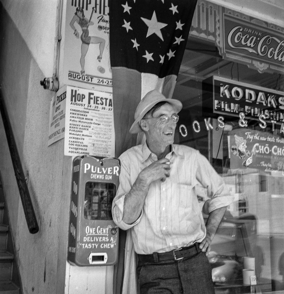 Independence man: A resident of Polk County, Oregon, hangs out on Main Street in the town of Independence. Dorothea Lange photographed the hop harvest in the area in August 1939. Near the man is a sign advertising the annual four-day “Hop Fiesta,” including an amateur hour, street dance, acrobats, log rolling and aquaplaning. He stands next to a Pulver chewing gum dispenser; for one cent, “delivers a tasty chew.”