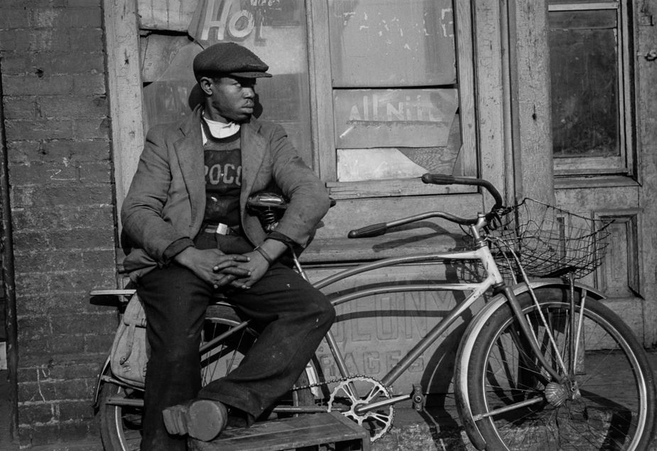 Young man and bicycle: A young Black man wearing a newsie cap sits with his bicycle outside a boarded-up building in Chicago’s “Black Belt.” The bike includes swept-back handlebars, a basket on the front and what looks like a canvas pannier on the back. Edwin Rosskam made the photograph in July 1941 while on assignment in Chicago.