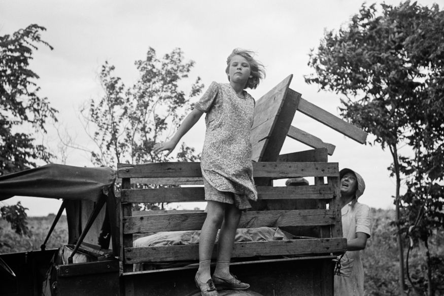 Migrant girl on truck: A girl “catches the wind” as her parents lift a table onto their truck before they leave Muskogee, Oklahoma, for California. Russell Lee photographed this family’s part in the “Okie” migration in July 1939.