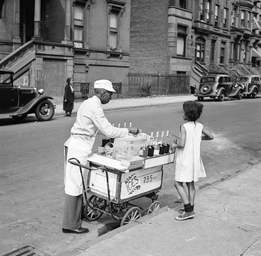 Ices: A young Black girl waits for a street vendor to make her an ice on a Harlem street in the summer of 1938. The vendor offers a selection of flavorings and three sizes, priced at 2, 3 and 5 cents. The vendor’s cart appears to have been converted from a baby carriage.