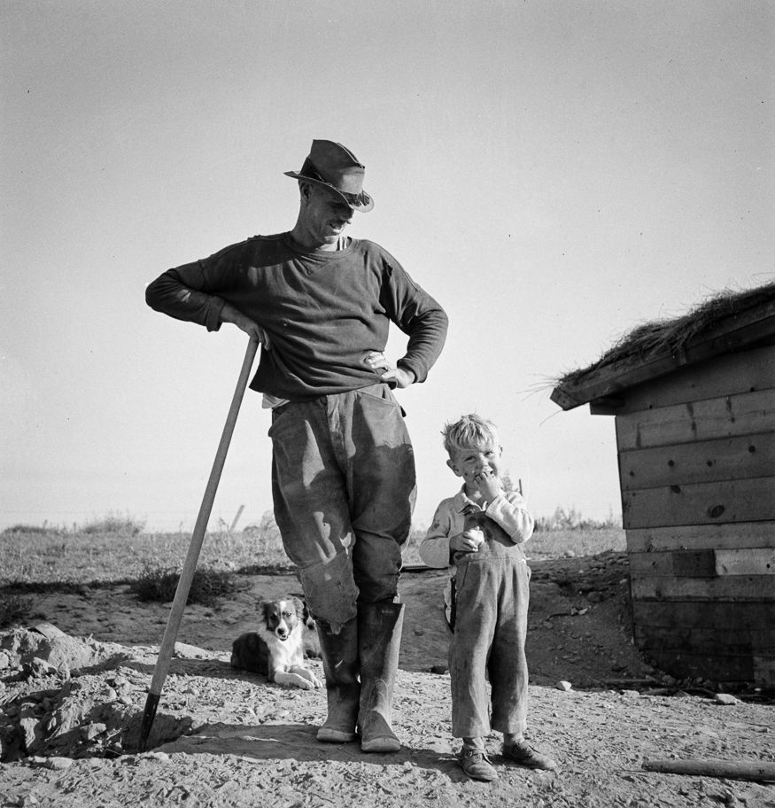 Father, son and dogs: In Malheur County, Oregon, on the eastern side of the state bordering Idaho, Dorothea Lange encountered Mr. Dougherty, who leans on a shovel, looking lovingly at his smiling young son, while two dogs rest nearby. The farmer, his wife and five children lived in a basement house - partially underground - and purchased their land with a loan from the Farm Security Administration. The U.S. Bureau of Reclamation built irrigation projects in the county to enable productive farming.