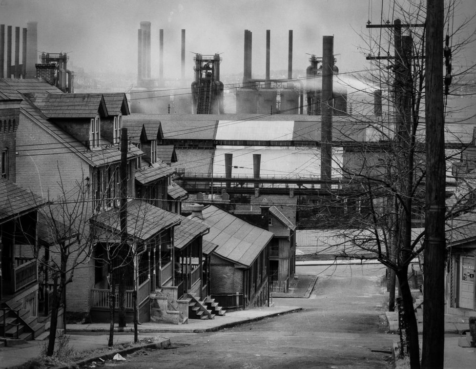 Steel mill neighborhood: Houses, leafless trees and electric poles descend a steep street in Bethlehem, Pennsylvania, leading to the stacks and buildings of the Bethlehem Steel Co. plant. Smoke and haze shroud the steel mill in the photograph, made by Walker Evans in November 1935.