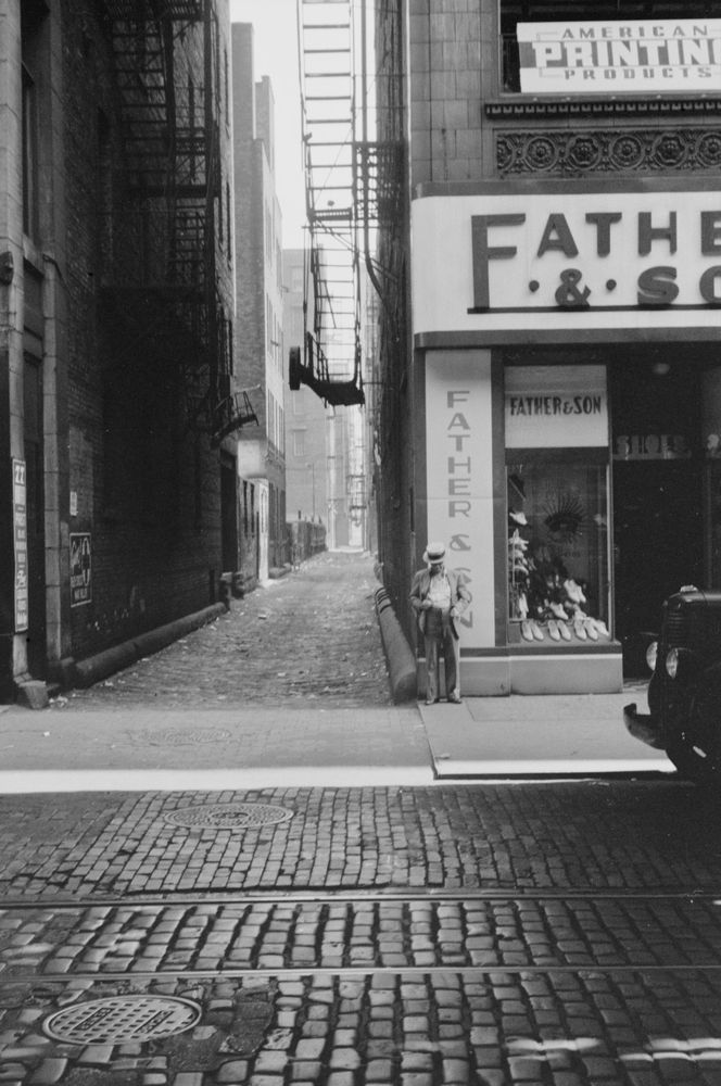 Waiting by the alley: John Vachon, in the summer of 1940, captured a man with a straw hat standing along a cobblestone and brick street in Chicago, Illinois. The Father & Son clothing store where he stands borders a long alley with prominent fire escapes.