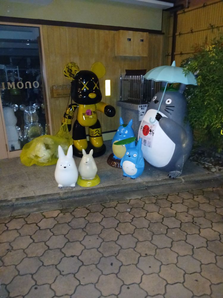 Statues of various ghiblo characters standing in a dingy corner with a giant black and yellow dead rat statue holding a big trashbags. They are likely scavenging for food. I guess the city encroached on the natural Totoro habitat.