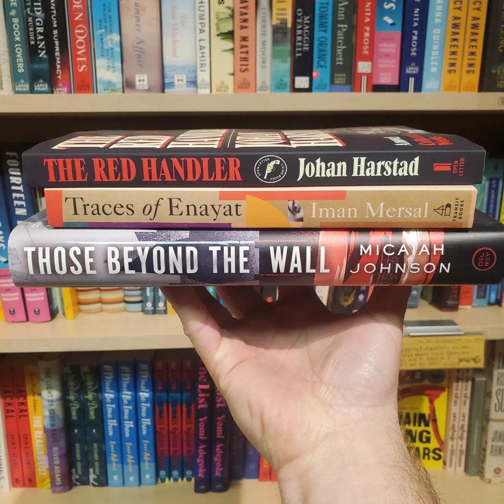 A hand holding aloft a stack of books containing Those Beyond the Wall, Traces of Enayat and The Red Handler with filled bookshelves in the background