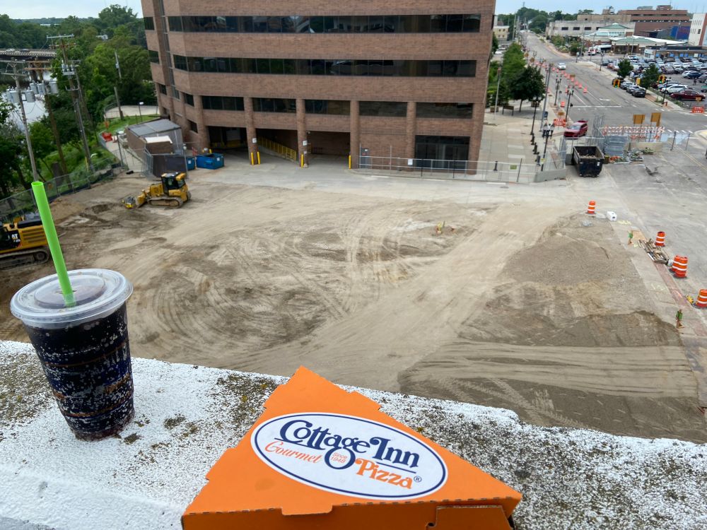 Single slice pizza box and a pop (soda) on a parking lot ledge overlooking a construction site for a skyscraper.
