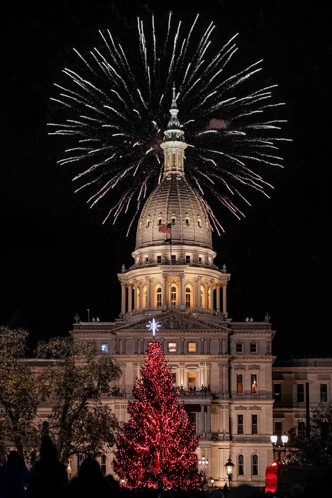 Image shows Michigan state Capitol building in dark with fireworks at the dome and a large Christmas tree in front covered in red lights and a white star.