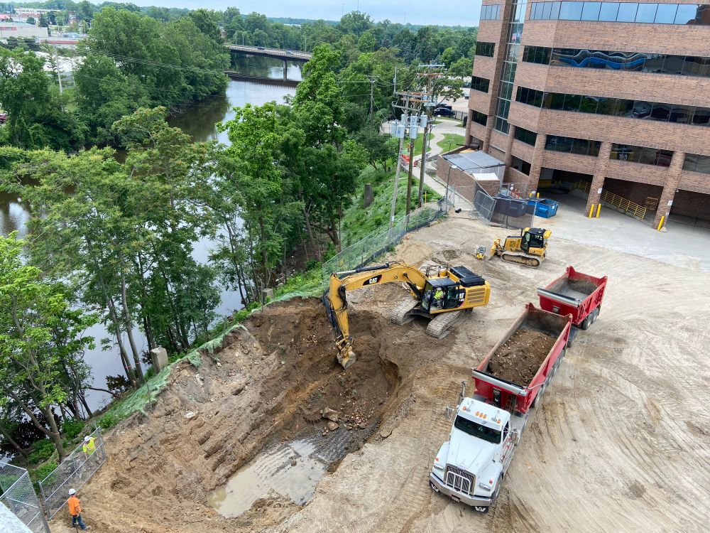 From a 5 story parking lot ledge overlooking a construction site for a skyscraper as heavy equipment digs next to a river.