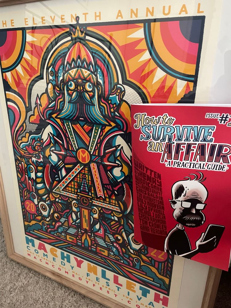 A poster for Machynlleth Comedy Festival 2022 in a wooden frame. The poster is designed by Drew Millward and is fun and colourful with the Mach clock tower as a centre point. To one side of the poster is a comic called “How to Survive an Affair: A Practical Guide”. It is written by Henry Widdicombe and illustrated by many excellent artists. The front of the comic has a red background and features an illustration of Henry looking at a phone, as well as the title and list of illustrators. 