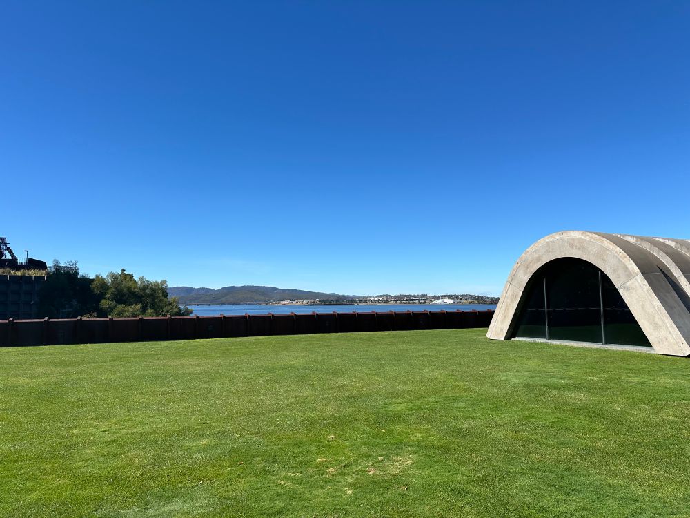 Photo from the natural lawned rooftop of Faro restaurant bar at MONA in Berriedale Tasmania. The Bowen Bridge can be seen in the far distance along with silhouette of natural bushland hills. The edge of the roof top has been fenced in architectural Corten steel that takes on a rich rusty  brown hue.  The parabolic concrete roofline of the Faro building juts out from the lawn like it is poking up its head. The sky is a rich flat blue colour devoid of clouds. The photo represents a perfect Hobart day, of moderate temperature, zero wind and calm beauty. Truly wonderful. 