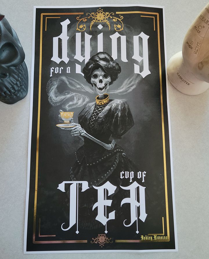 A print of a black and white painting of a skeleton with a Victorian-style updo and dress. She is holding a cup of tea, from which steam swirls back and around to frame the head. There are gold accents on the dress and teacup as well as gilded decorative framing elements.
The text says: “Dying for a cup of tea.”
