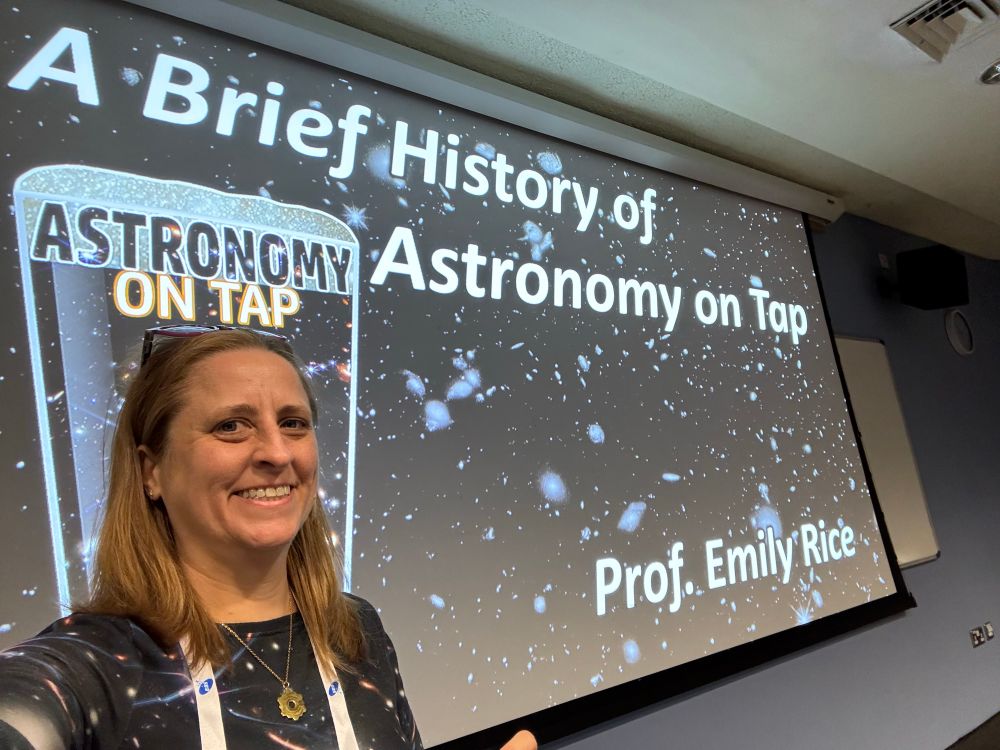 Selfie of Emily in front a large screen showing the opening slide of “A Brief History of Astronomy on Tap” with the AoT pint glsss logo and a deep field of galaxies background image 