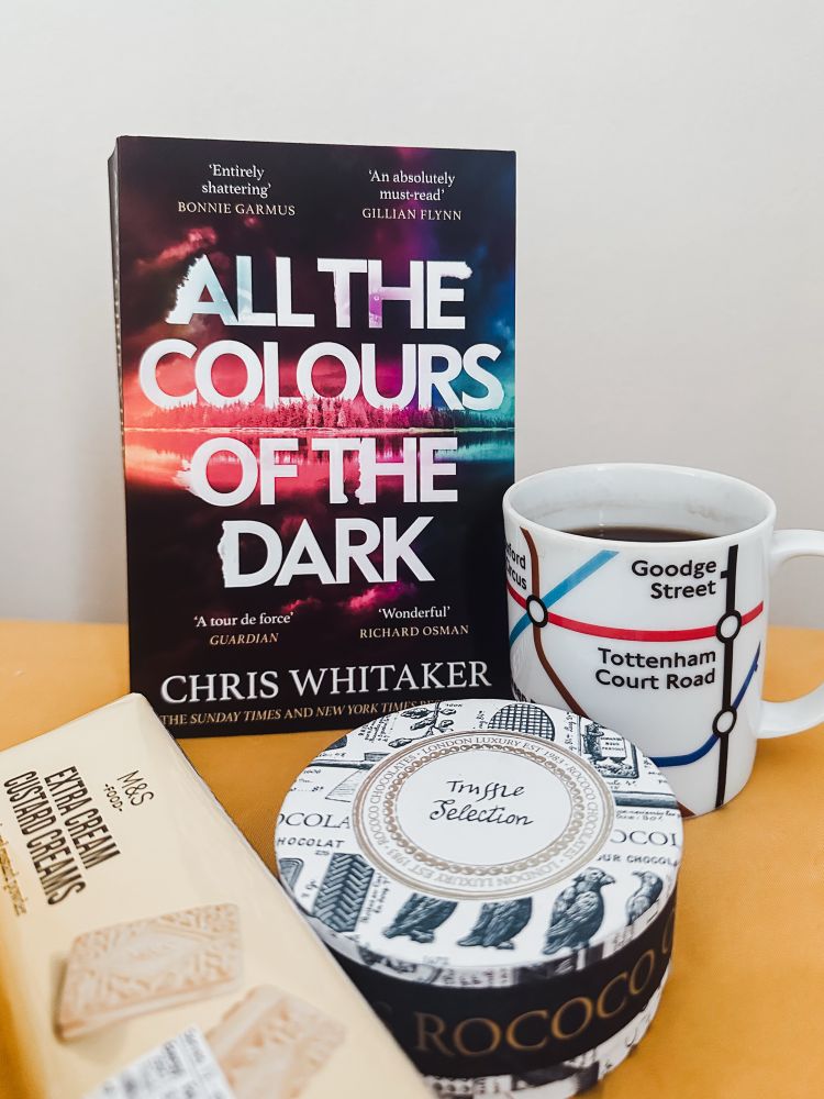 All Colours of the Dark book, black Americano in a London Underground mug, box of rococo truffles and a pack of M&S extra cream custard creams on a table