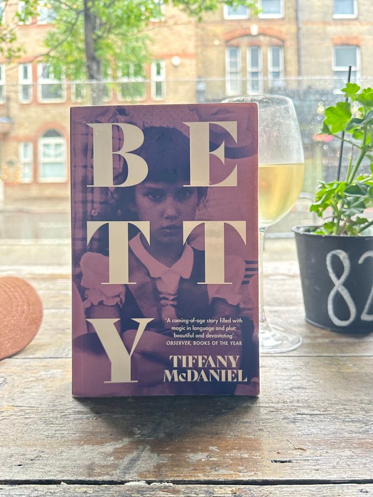 Paperback version of Tiffany McDaniel’s Betty per ch ed on a gastropub, glass of white wine and a potted plant with number 82. Block of flats and offices in the background. 
