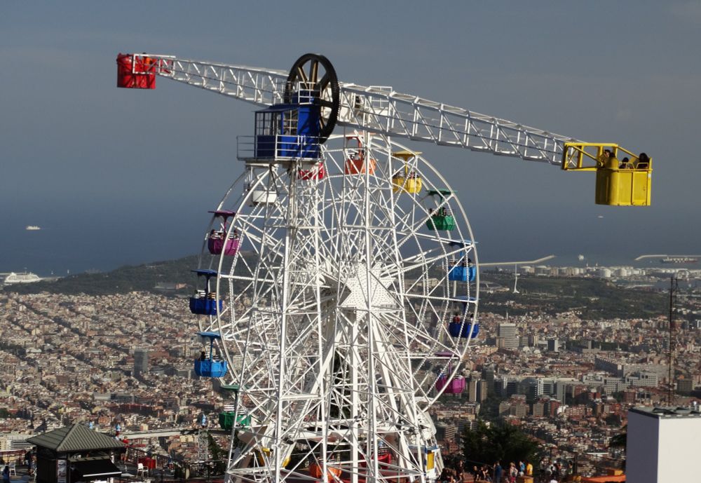 A white Ferris Wheel with multicoloured chairs and a bucket swing ride in the foreground of a view of the sea, Barcelona and Montjuic with mist in the distance.