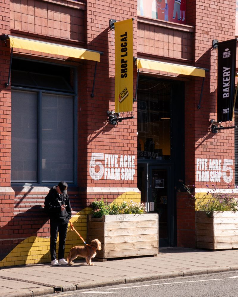 A man dressed in black stands by the wall of a red-brick shop called Five Acre Farm Shop, holding his dog’s leash.
The yellow awnings read SHOP LOCAL.
The morning light is sharp, cutting the scene in halves: routine and pause, city and silence.