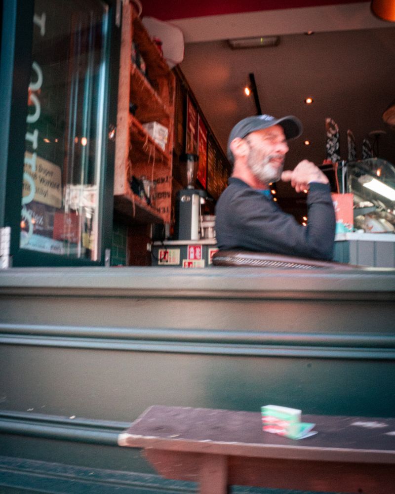Blurred shot of a man laughing inside a small café.
The green counter, red walls, and stray light outside form a frame of movement and warmth — the kind of imperfection that feels alive.