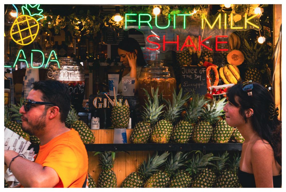 A neon-lit juice stall packed with pineapples and fruit. In the foreground, two passersby cross in opposite directions, one in a bright orange shirt, the other in a black strapless top, their faces frozen mid-motion. The scene feels like a slice of urban theater, with light, fruit, and people competing for attention.
