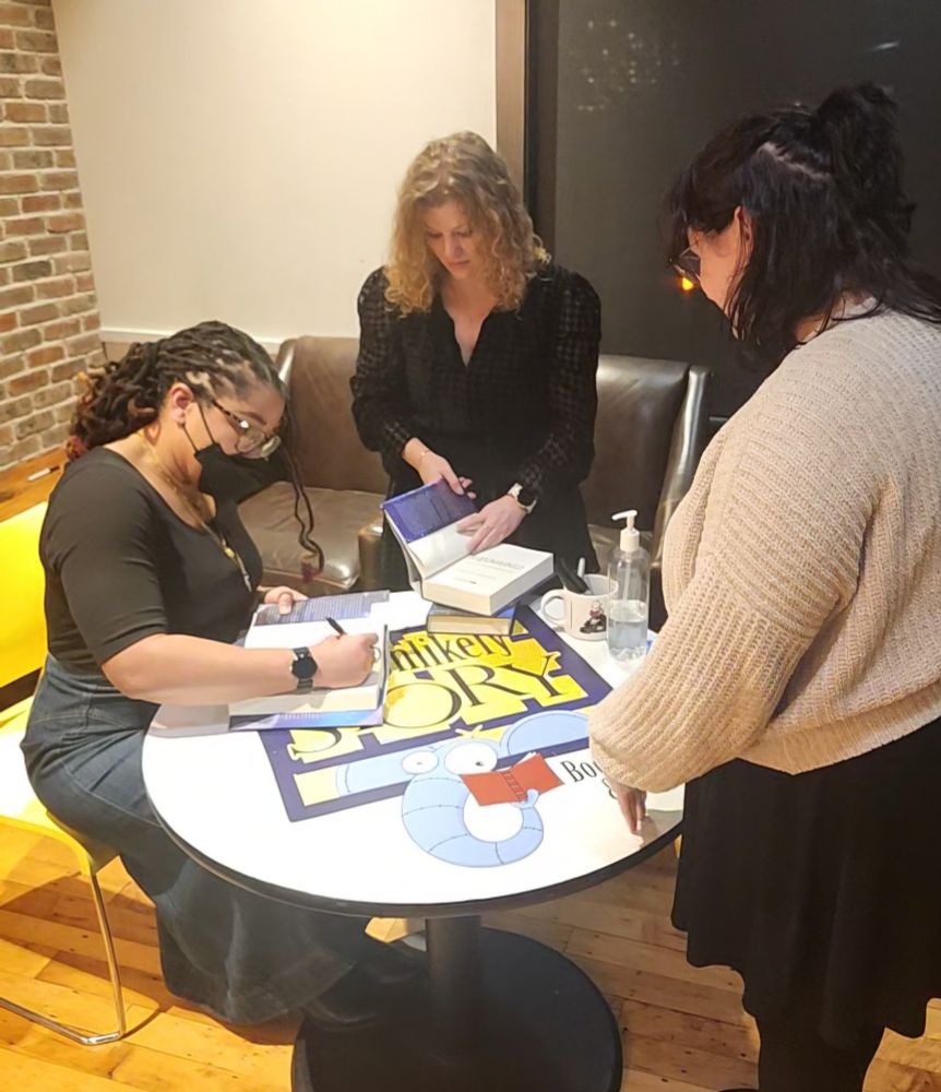 Three woman are around a table. Tracy Deonn is sitting, signing a book, with an assistant in the middle opening a book, and OP standing to the right