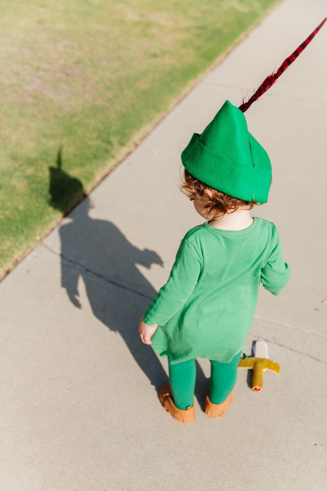 toddler dressed as peter pan standing on the sidewalk looking at his shadow 