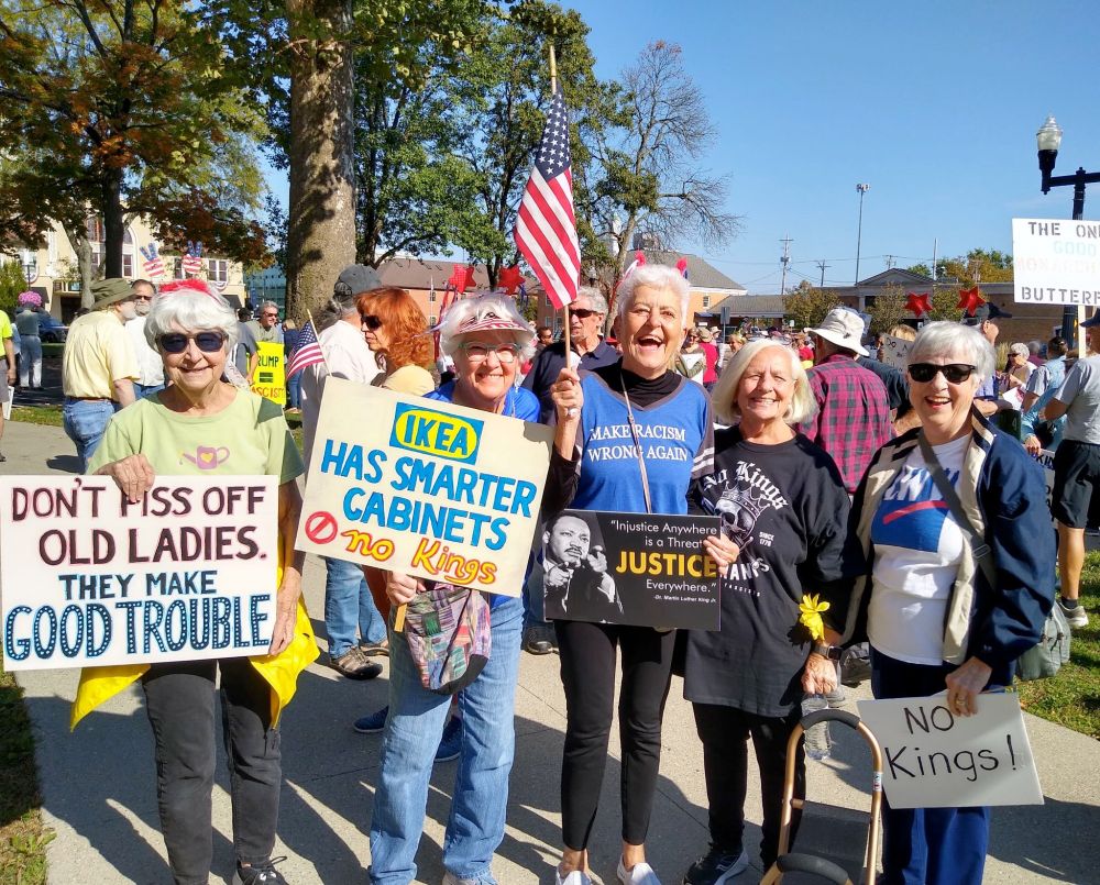 A photo of five smiling white-haired women, with a crowd in the background. Their signs read: "DON'T PISS OFF OLD LADIES. THEY MAKE GOOD TROUBLE"; "IKEA HAS SMARTER CABINETS 🚫 No Kings"; "Injustice Anywhere is a Threat to Justice Everywhere - Martin Luther King Jr."; and "NO Kings!" The woman in the center holds a US Flag. 