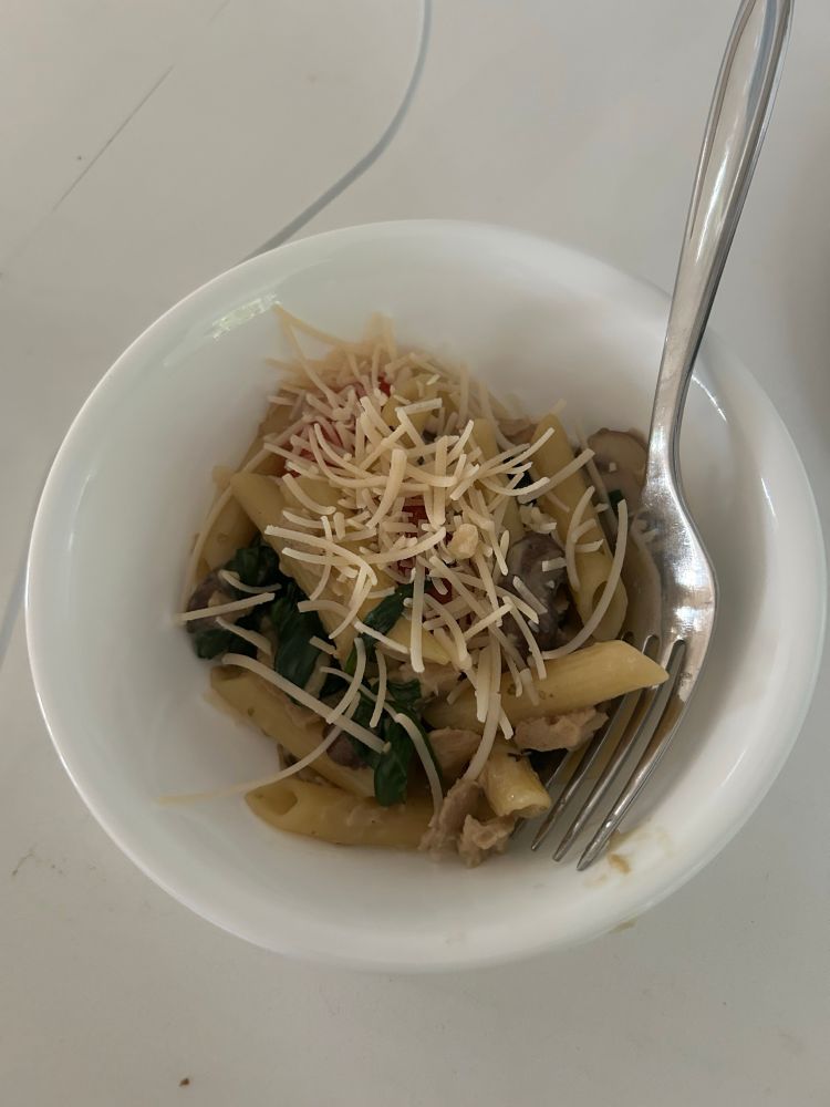A white bowl of vegan pasta with vegan cheese, spinach, mushrooms, and tomatoes.