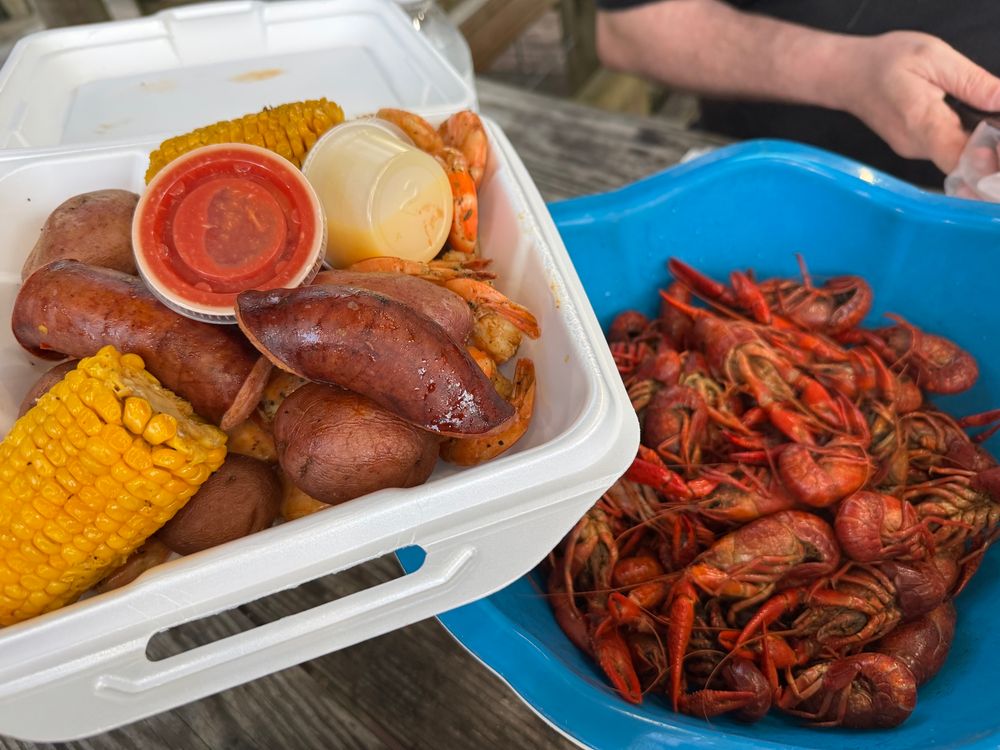A large white styrofoam container full of sausage, corn, potatoes, and shrimp held over a large blue bowl of cooked crawfish.