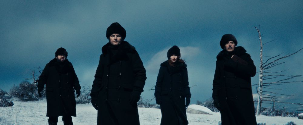 Four people stand in a snowy landscape against a dark teal sky. They are dressed identically in black winter coats and black knit caps, creating a stark silhouette effect. The figures are positioned in a loose line formation with the second person from the left appearing slightly more prominent. Bare winter tree branches are visible on the right side of the frame. The image has a cold, cinematic quality with a muted color palette dominated by blues and blacks.