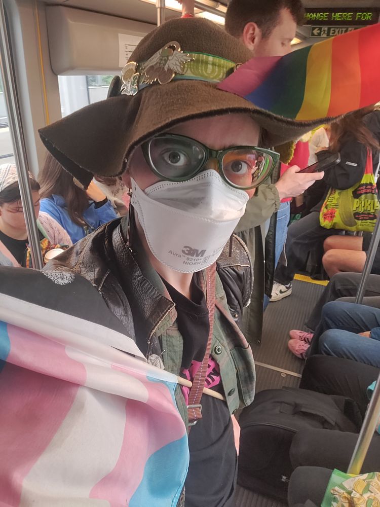 A cute white genderqueer person on a decently crowded green line train. They have a trans pride flag stuck in their purse strap and a progress pride flag in their hatband. They are wearing bright green glasses, amazing rainbow eye makeup, and a white n95 mask. 